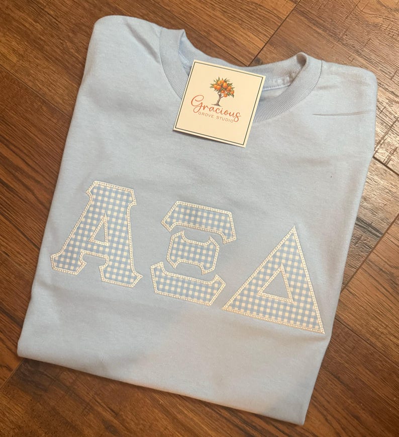 May include: Light blue t-shirt with the Greek letters ASA in white and light blue gingham fabric. The shirt has a tag that reads "Gracious Grove Studio". The shirt is folded on a wooden surface.