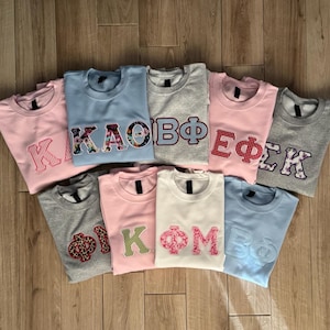 May include: A collection of nine crewneck sweatshirts in various colors including light blue, light pink, grey, and white. Each sweatshirt features large, fabric-appliqued Greek letters on the chest, showcasing diverse patterns like floral, plaid, and leopard print.