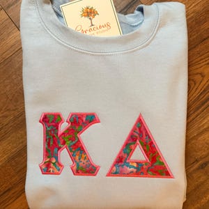 May include: A light blue sweatshirt with the embroidered letters "KA" in a colorful floral pattern. The sweatshirt has a crew neck and a tag that reads "Gracious Love Studio".