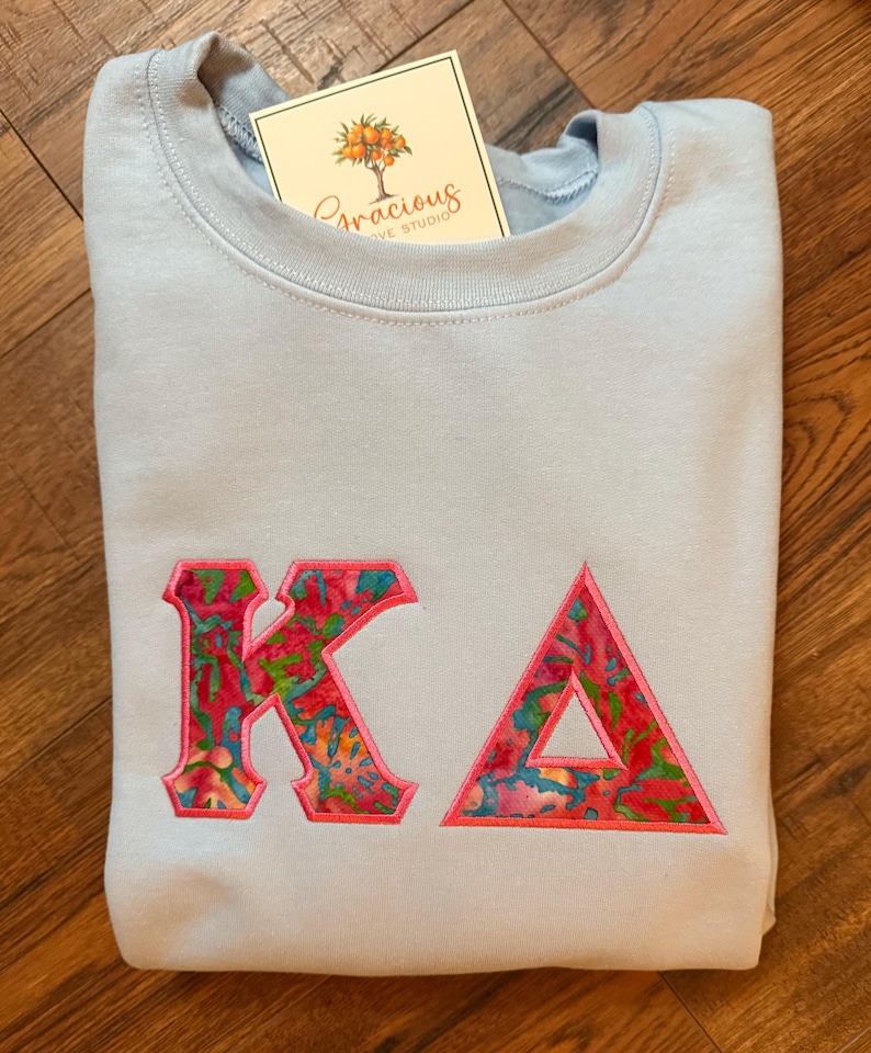 May include: A light blue sweatshirt with the embroidered letters "KA" in a colorful floral pattern. The sweatshirt has a crew neck and a tag that reads "Gracious Love Studio".