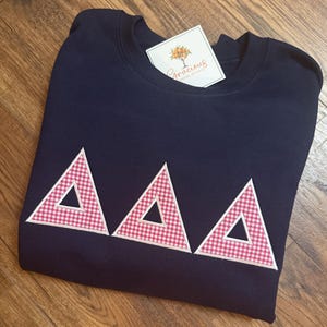 May include: Navy blue sweatshirt with three red and white gingham triangles. The triangles are outlined in white. A tag with the text "Gracious Grove Studio" is attached to the sweatshirt.