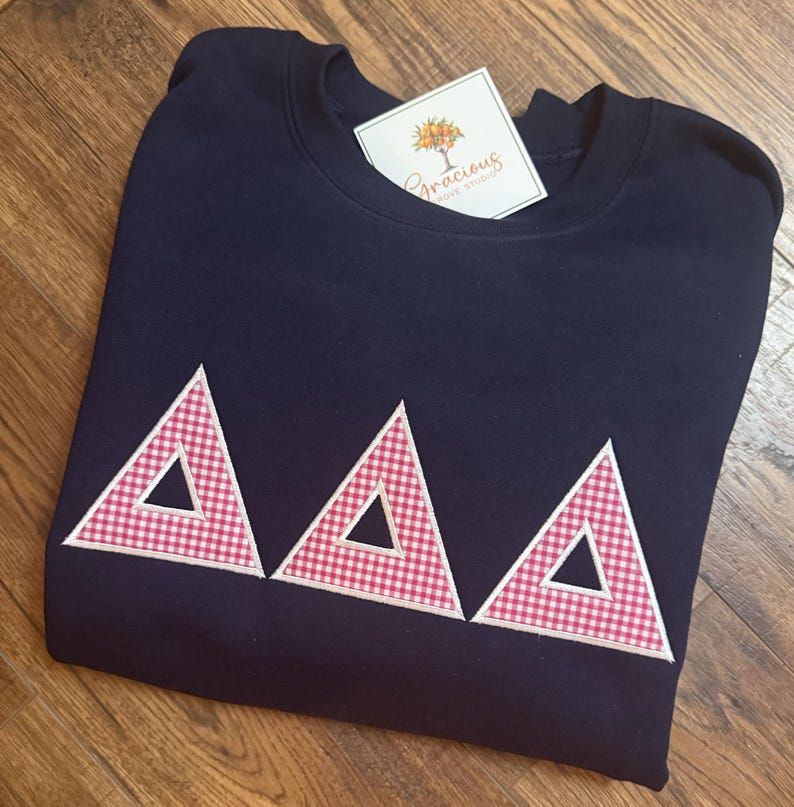 May include: Navy blue sweatshirt with three red and white gingham triangles. The triangles are outlined in white. A tag with the text "Gracious Grove Studio" is attached to the sweatshirt.