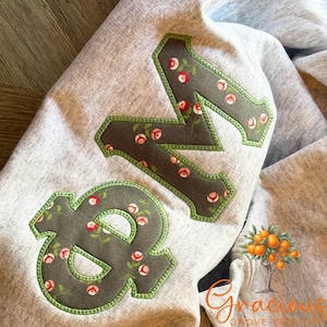 May include: Gray sweatshirt with large, decorative letters "&N" appliqued on the front. The letters are a dark gray with a floral pattern and a green border. The sweatshirt is folded.