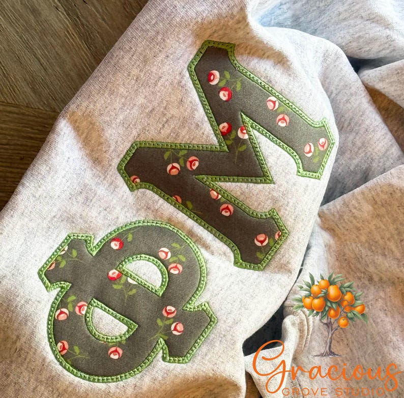 May include: Gray sweatshirt with large, decorative letters "&N" appliqued on the front. The letters are a dark gray with a floral pattern and a green border. The sweatshirt is folded.