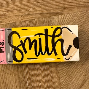 May include: A yellow and brown wooden sign shaped like a pencil with the text "Miss. Smith" on it.