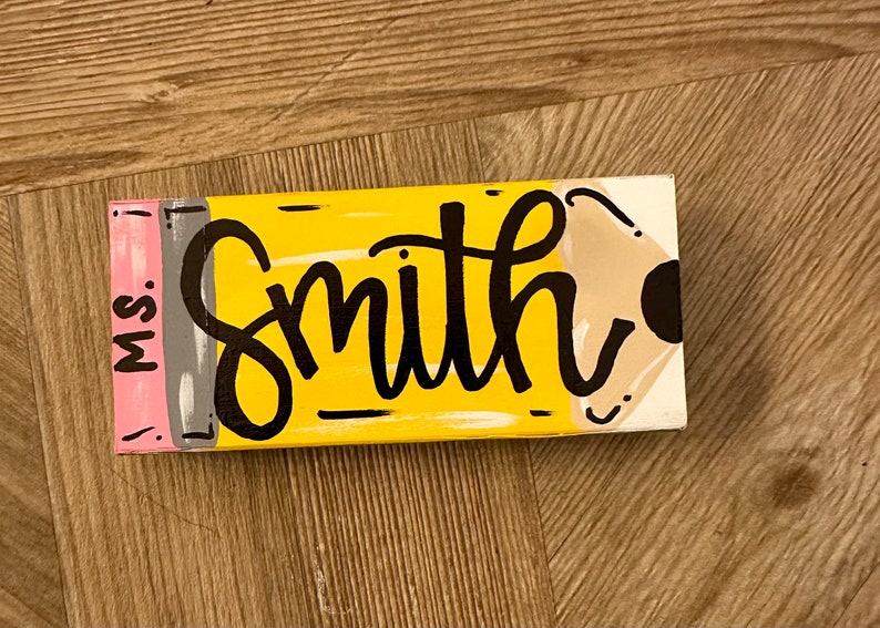 May include: A yellow and brown wooden sign shaped like a pencil with the text "Miss. Smith" on it.
