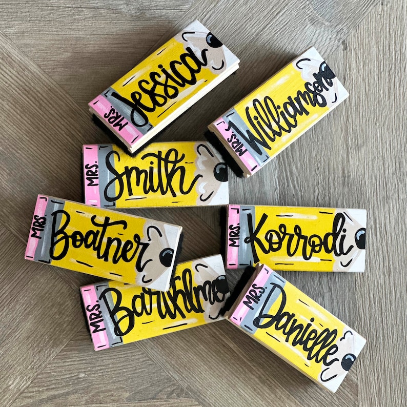 May include: Eight yellow pencil-shaped wooden erasers with pink eraser tips. Each eraser has a black handwritten name on it, including "Mrs. Jessica", "Mrs. Smith", "Mrs. Williamson", "Mrs. Boatner", "Mrs. Korrodi", "Mrs. Baruklm", and "Mrs. Danielle".