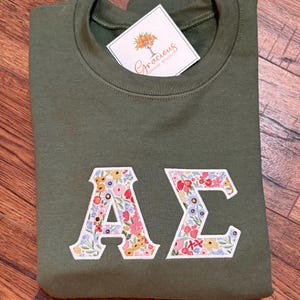 May include: An olive green sweatshirt with floral embroidered Greek letters, "A" and "&Sigma;". A white tag with the text "Gracious Grove Studio" is attached. The sweatshirt is folded on a wooden surface.