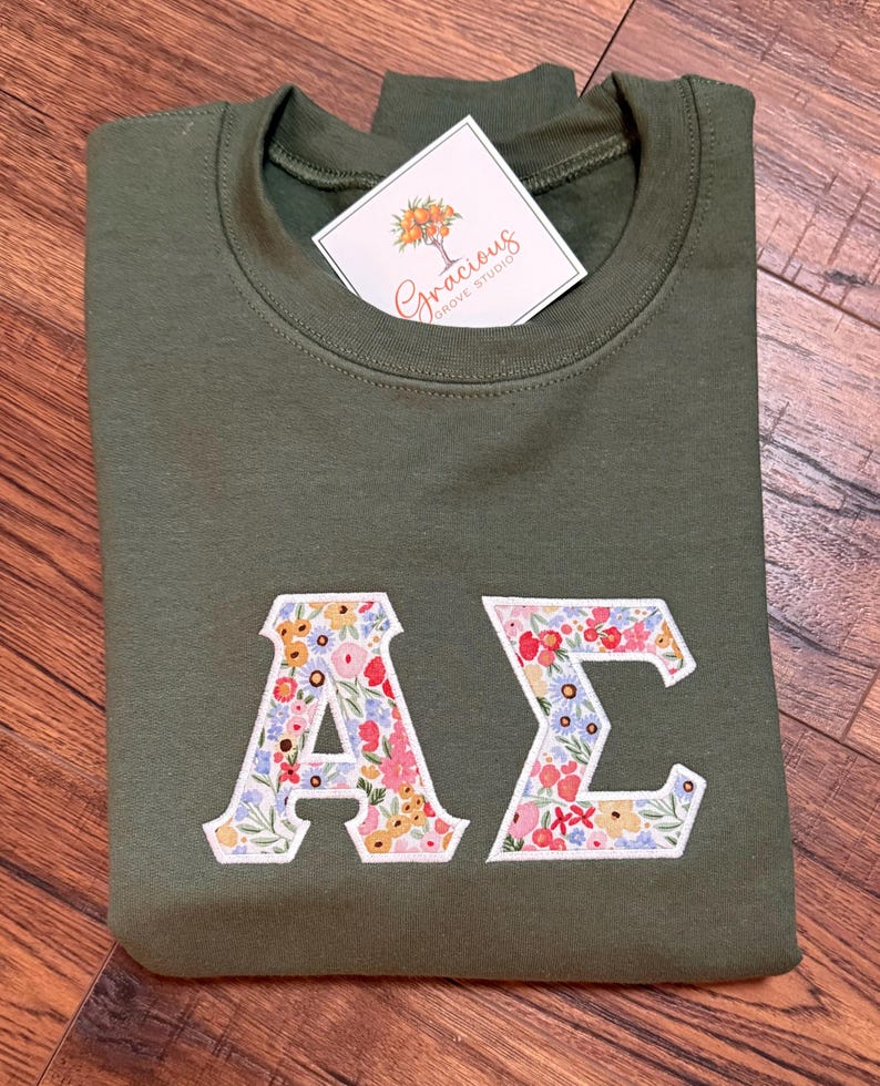 May include: An olive green sweatshirt with floral embroidered Greek letters, "A" and "&Sigma;". A white tag with the text "Gracious Grove Studio" is attached. The sweatshirt is folded on a wooden surface.