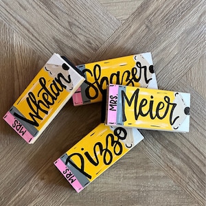 May include: Four yellow pencil-shaped wooden signs with black lettering. The signs read "Mrs. Whelan", "Mrs. Slazer", "Mrs. Meier", and "Mrs. Pizzo".