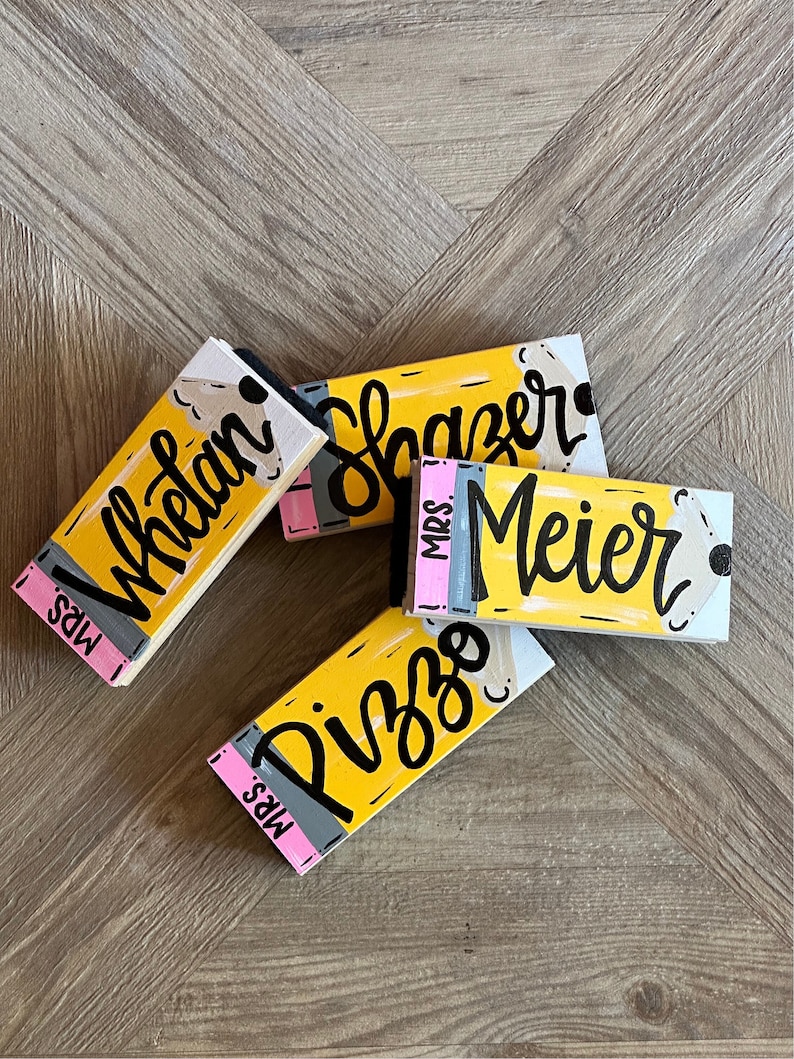 May include: Four yellow pencil-shaped wooden signs with black lettering. The signs read "Mrs. Whelan", "Mrs. Slazer", "Mrs. Meier", and "Mrs. Pizzo".