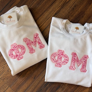 May include: Two white sweatshirts with the Greek letters &Phi; and M in pink, patterned fabric. Each sweatshirt has a tag with a tree logo and the word "Gracious" on it. The sweatshirts are folded on a wooden surface.