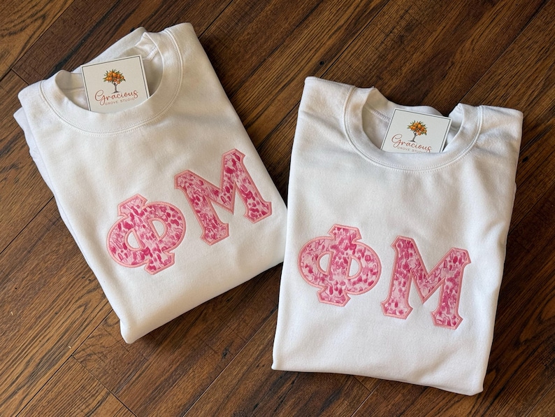 May include: Two white sweatshirts with the Greek letters &Phi; and M in pink, patterned fabric. Each sweatshirt has a tag with a tree logo and the word "Gracious" on it. The sweatshirts are folded on a wooden surface.