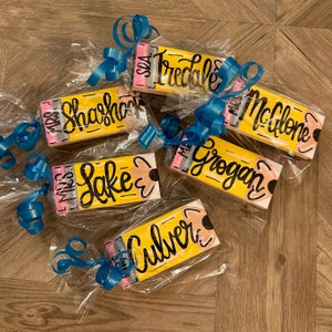May include: Six yellow pencil-shaped wooden gift tags with black and white lettering. The tags are wrapped in clear cellophane and tied with blue ribbon. The names on the tags are "Miss Shahshock", "Miss Tredale", "Miss McGlone", "Miss Grogan", "Miss Lake", and "Miss Culver".