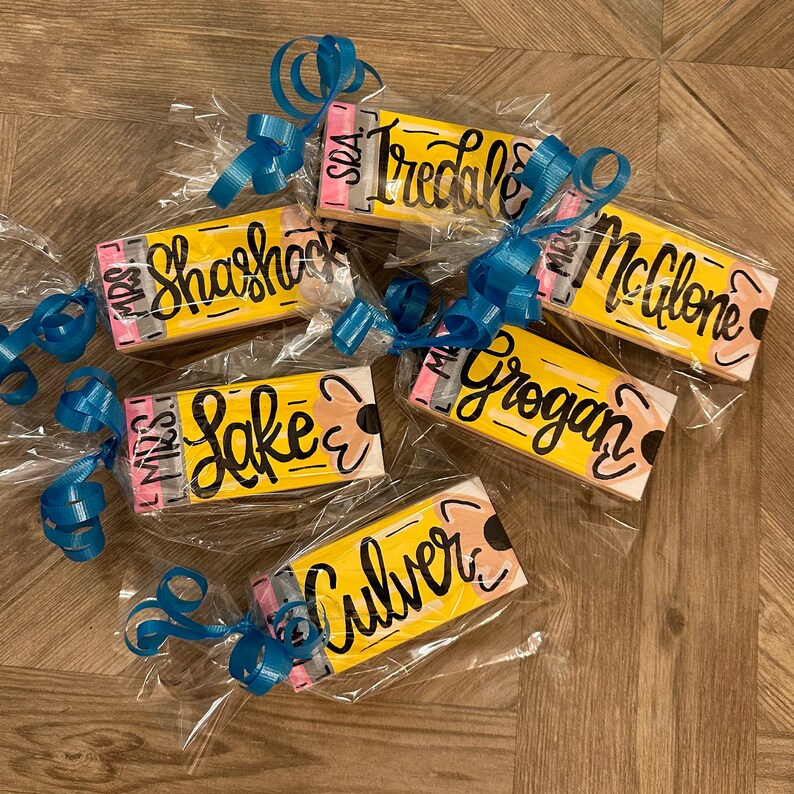 May include: Six yellow pencil-shaped wooden gift tags with black and white lettering. The tags are wrapped in clear cellophane and tied with blue ribbon. The names on the tags are "Miss Shahshock", "Miss Tredale", "Miss McGlone", "Miss Grogan", "Miss Lake", and "Miss Culver".