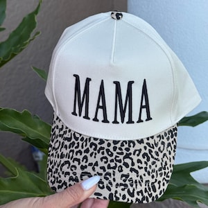 May include: White baseball cap with the word "MAMA" embroidered in black. The brim features a leopard print design. The cap is a stylish accessory.