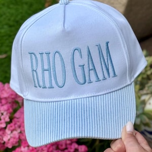 May include: A white baseball cap with the embroidered text "RHO GAM" in light blue. The brim features a light blue and white striped pattern. The cap is held by a person in front of a background of pink flowers.