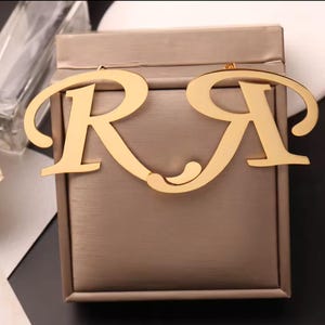 May include: Gold-colored earrings in the shape of the letters "R" and "A". The earrings are displayed on a textured, taupe-colored box. The letters are connected by a small, curved design.