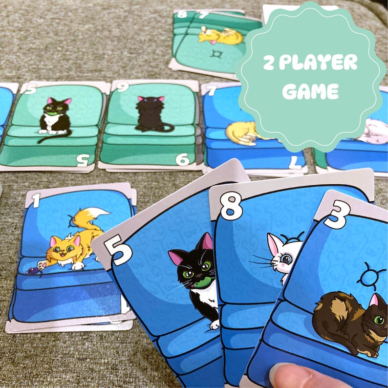 May include: A 2 player game with cards featuring cats and couches. The cards have numbers on them and show different colored cats sitting on blue couches. The text on the card reads "2 Player Game".