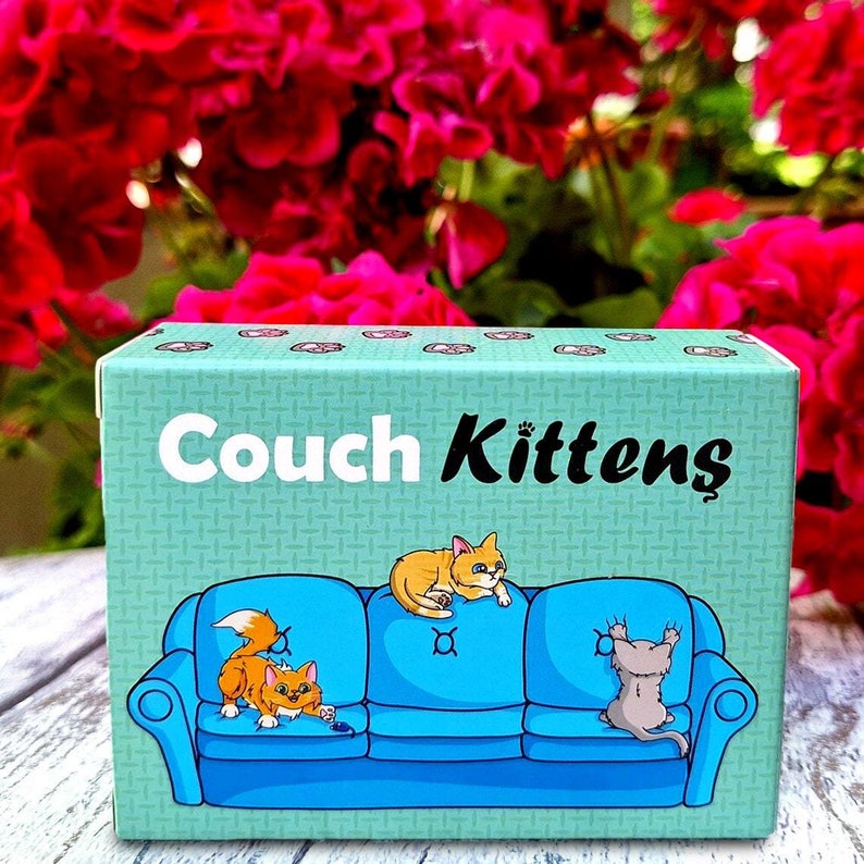 May include: A blue and green box with the text "Couch Kittens" on the front. The box features an illustration of a blue couch with three cats sitting on it. The cats are a yellow tabby, a white and gray tabby, and a brown tabby.