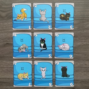 May include: Nine blue cards with white numbers and white outlines. Each card features a different cat illustration in a blue armchair. The cards are numbered 1 through 9.