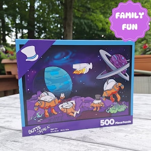 May include: A 500-piece puzzle box featuring a cartoon illustration of four astronauts in space. The box is blue and purple with the words "BUTTS in SPACE" and "FAMILY FUN" on it. The puzzle is 20.5 x 15 inches.