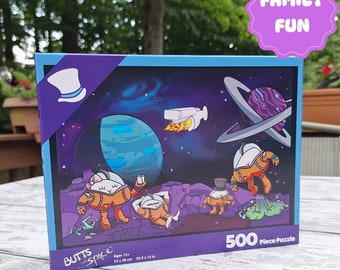 May include: A 500-piece puzzle box featuring a cartoon illustration of four astronauts in space. The box is blue and purple with the words "BUTTS in SPACE" and "FAMILY FUN" on it. The puzzle is 20.5 x 15 inches.