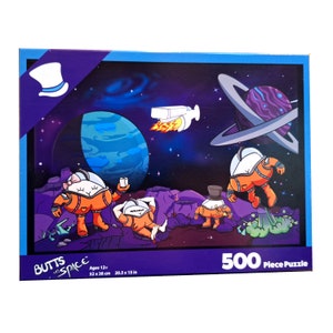 May include: A 500 piece jigsaw puzzle featuring a cartoon illustration of astronauts in space. The puzzle box has a purple background with the words "Butts in Space" and a cartoon top hat. The puzzle is for ages 12 and up and measures 20.5 x 15 inches.