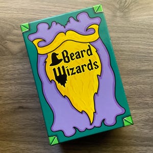 May include: A rectangular box with a teal green background and a cartoon illustration of a yellow beard with a black hat and the words "Beard Wizards" in black and yellow. The box has purple and green accents.