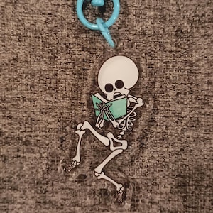 May include: A clear acrylic keychain featuring a cartoon skeleton reading a teal book. The skeleton is white with black eye sockets and is attached to a light blue key ring. The background is a textured gray fabric.