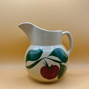 May include: A cream-colored ceramic pitcher with a green and red apple design. The pitcher has a round body and a handle.