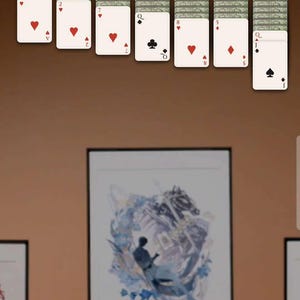 May include: A mobile game of Chibi Solitaire is displayed on a phone screen. The game shows a deck of cards and a framed artwork on a table. The game's interface includes the text "CHIBI SOLITAIRE".