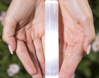 May include: A long, white selenite wand, held between two hands. The wand is cylindrical with a rounded tip and a textured surface. The background is a soft green with blurred foliage.