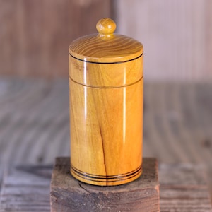 May include: A cylindrical wooden container with a lid. The container is a light golden color with visible wood grain. It has a small, round knob on top and black accent lines. The container is sitting on a wooden block.