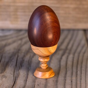 May include: A dark brown wooden egg sits on a light brown wooden egg cup. The egg cup has a round base and a stem with a decorative design.