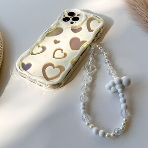 May include: A white phone case with a gold heart pattern and the word "Love" printed on it. The case is attached to a white beaded phone strap with a white cloud charm.
