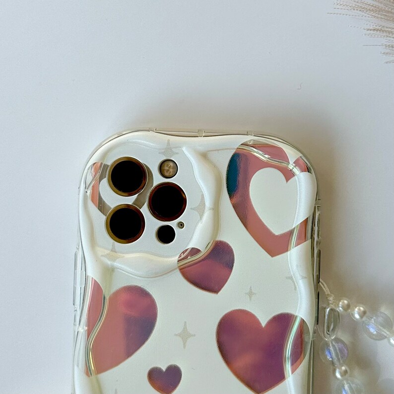 May include: A clear phone case with a white background and a pink holographic heart pattern. The case has a raised lip around the camera lens and a clear strap.