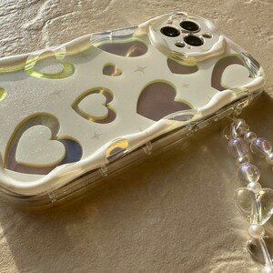 May include: A clear phone case with a white background and a pattern of hearts. The case has a beaded strap attached to it.