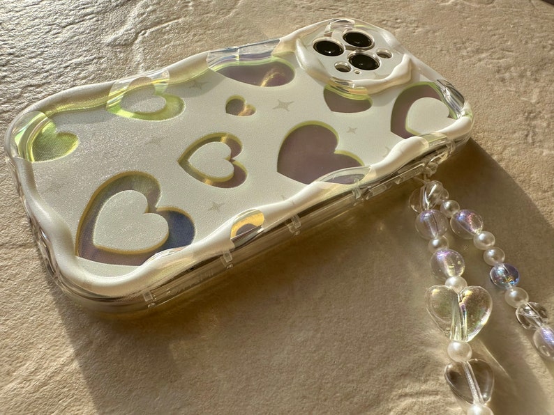May include: A clear phone case with a white background and a pattern of hearts. The case has a beaded strap attached to it.