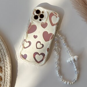 May include: A clear phone case with a pink and white heart pattern and the word "Love" printed on it. The case is attached to a white beaded phone strap with a cloud-shaped charm.