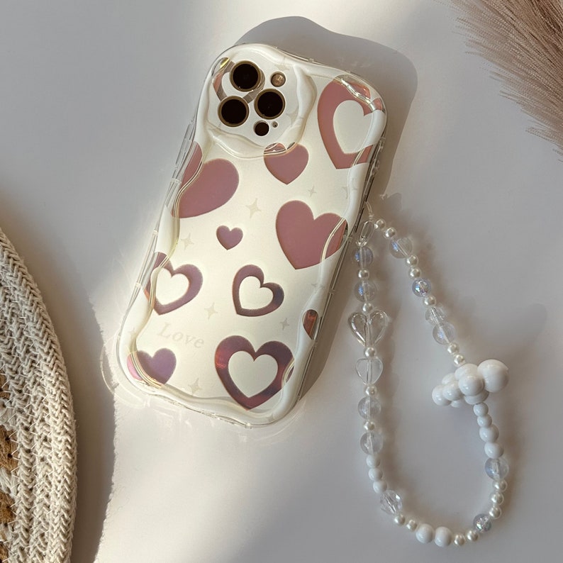 May include: A clear phone case with a pink and white heart pattern and the word "Love" printed on it. The case is attached to a white beaded phone strap with a cloud-shaped charm.