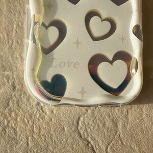 May include: A clear phone case with a white background and a holographic design of hearts and stars. The case has the word "Love" printed on it.