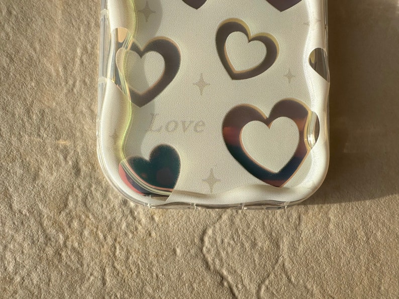 May include: A clear phone case with a white background and a holographic design of hearts and stars. The case has the word "Love" printed on it.