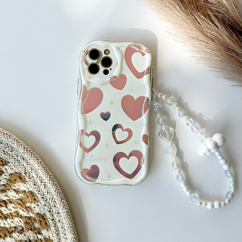 May include: A clear phone case with a pink and white heart pattern and the word "Love" printed on it. The case has a white beaded strap attached.