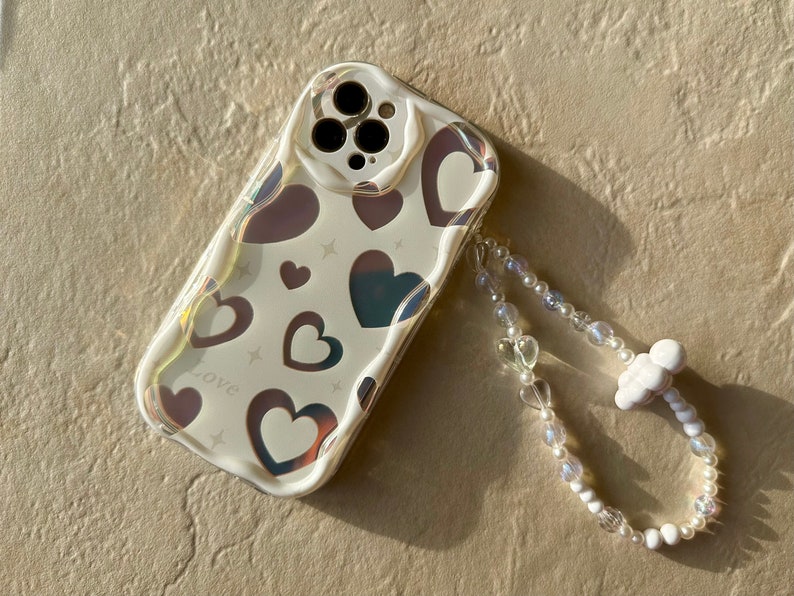 May include: A white phone case with a holographic heart pattern and the word "love" printed on it. The case has a clear camera lens and comes with a white beaded phone strap.