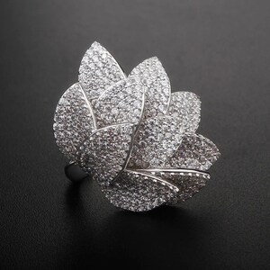 May include: A silver ring with a large, multi-layered flower design. The flower is made up of many small, clear stones.