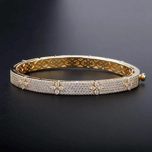 May include: A gold-colored bangle bracelet with a floral design. The bracelet is encrusted with small, clear gemstones and features a decorative pattern on the inner band. The clasp is visible on the side. The bracelet is displayed on a black surface.
