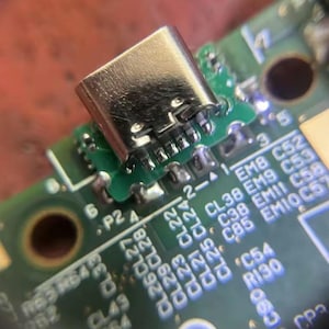 May include: Close-up of a USB-C connector soldered onto a green circuit board. The connector is silver and has multiple pins. The board has white text and numbers, including "CL38" and "C52".