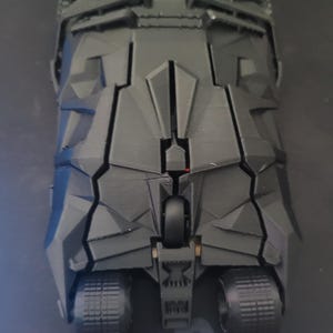 May include: A black, angular model of a vehicle with large, textured tires. The vehicle has a geometric design with multiple panels and a central wheel. The model is likely a replica of a fictional vehicle.