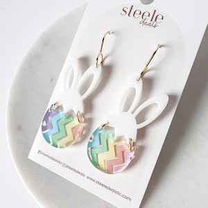 May include: A pair of gold hoop earrings with white bunny shaped charms and colorful chevron patterned Easter egg charms. The earrings are displayed on a white card with the text "steele deals".